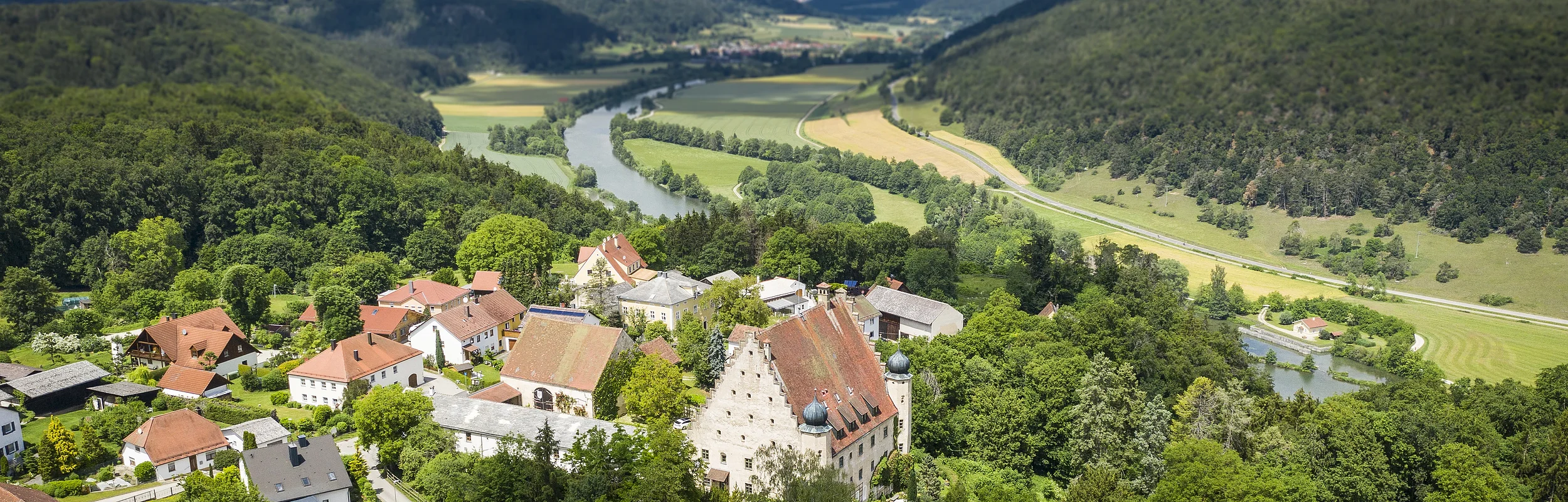 Luftaufnahme eines Dorfes mit Fluss, umgeben von bewaldeten Hügeln und Feldern unter bewölktem Himmel.