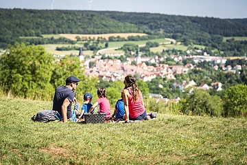 Familie mit drei Kindern sitzt auf einer Wiese mit Picknickkorb und Blick auf eine Stadt im Tal.