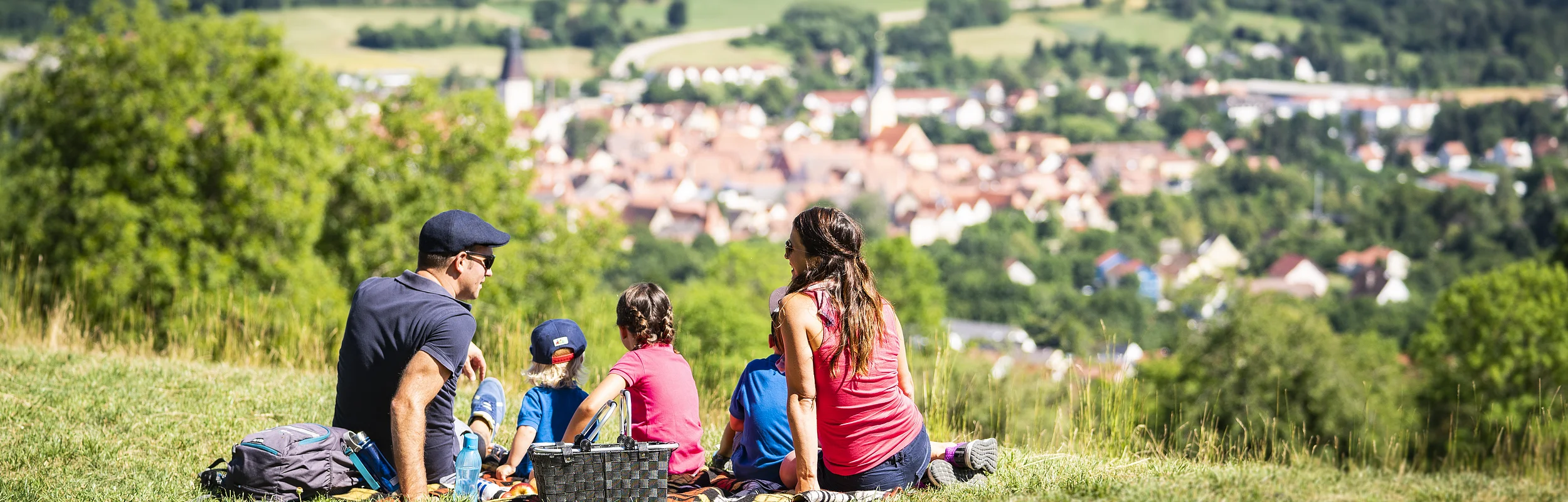 Familie mit drei Kindern sitzt auf einer Wiese mit Picknickkorb und Blick auf eine Stadt im Tal.