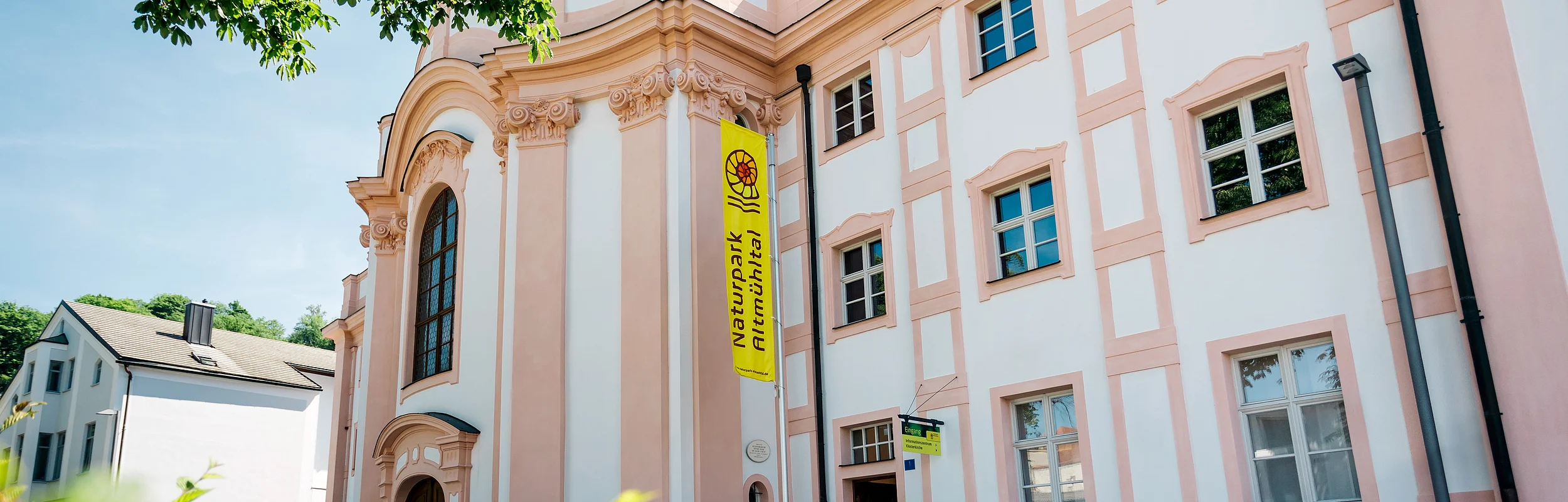 Der Eingangsbereich des Informationszentrum Naturpark Altmühltal Eichstätt ist vom Gehsteig schräg gegenüber zu sehen. Die gelbe Fahne und die Eingangstüre befinden sich im Mittelpunkt.