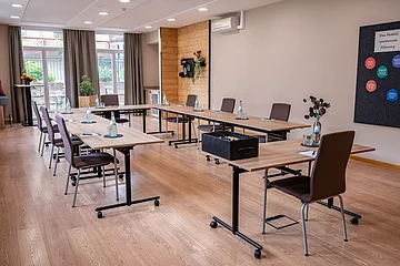 Konferenzraum mit U-förmig angeordneten Tischen, Stühlen, Wasserflaschen und Notizblöcken, Fenster mit Vorhängen.
